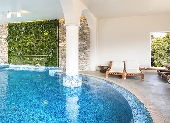 Innenpool mit blauen Mosaikfliesen, Wasserfall und Liegestühlen vor einer Steinwand mit Pflanzen.