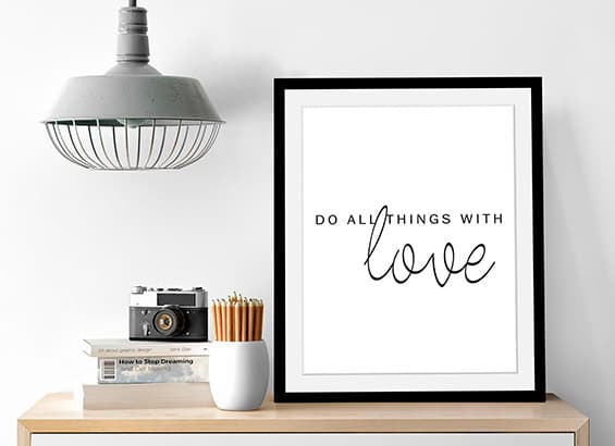 Affiche « Do all things with love » encadrée, posée sur une étagère avec appareil photo et pot de crayons
