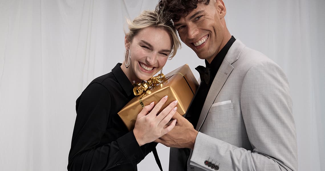 WEIHNACHTEN – Zwei Personen in festlicher Kleidung halten ein goldfarbenes Geschenk mit Schleife