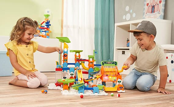 Deux enfants jouent avec un circuit de billes coloré sur le sol d'une chambre.