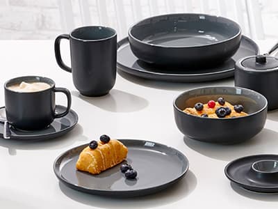 Schwarzes Geschirr-Set mit Tassen, Tellern und Schüsseln, serviert mit Croissant und Beeren auf einem Tisch.