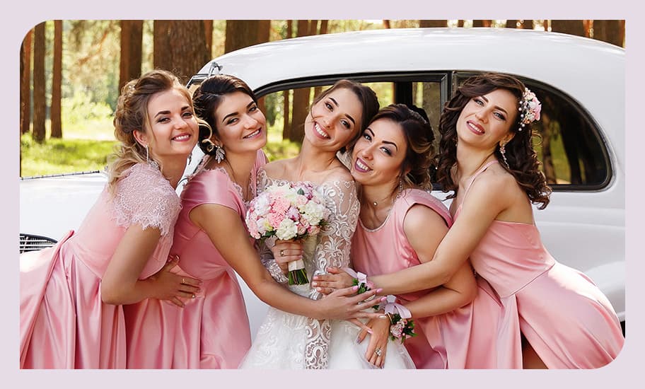 Braut in weißem Kleid posiert mit vier Brautjungfern in rosa Kleidern vor einem weißen Oldtimer-Hochzeitsauto