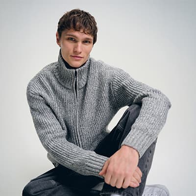 Junger Mann im grauen Strickpullover mit hohem Kragen sitzt entspannt auf dem Boden und blickt in die Kamera