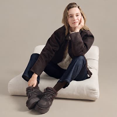 Person sitzt mit überschlagenen Beinen am Boden, trägt braune Jacke, blaue Jeans, weiße Socken und schwarze Schuhe
