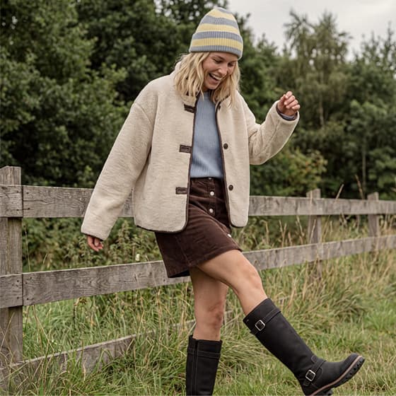 Frau im Herbstoutfit hebt ein Bein neben Holzzaun auf Wiese, trägt Jacke, Rock, Mütze und hohe Stiefel