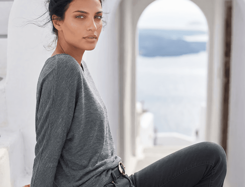 Frau im grauen Langarmshirt sitzt seitlich auf einer weißen Treppe mit Meerblick durch einen hellen Torbogen