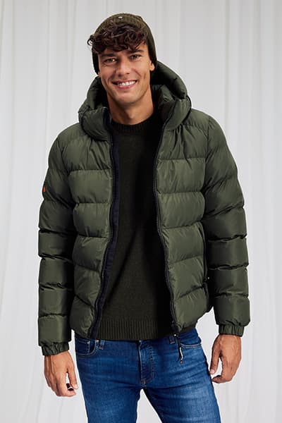 Mann mit lockigen Haaren trägt olivgrüne Steppjacke mit Kapuze, dunklen Pullover und blaue Jeans, steht frontal und lächelt