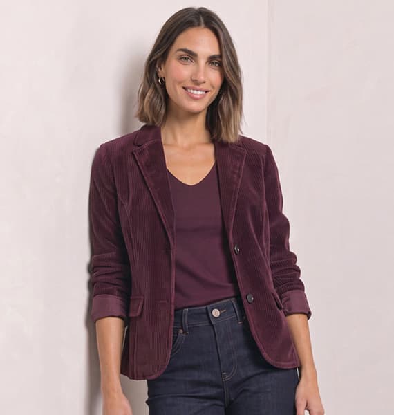 Frau mit schulterlangem Haar trägt bordeauxfarbenen Blazer über passendem Oberteil und dunkler Jeans, steht lächelnd vor hellem Hintergrund