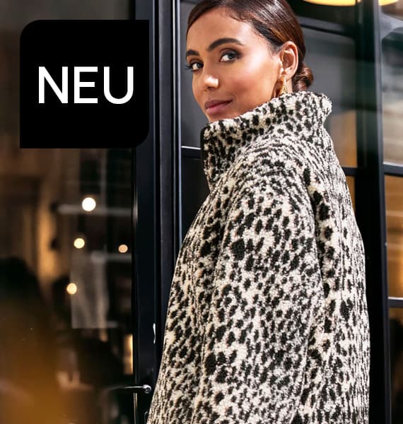 NEU – Frau mit lockigen Haaren trägt hellen Mantel und Sonnenbrille und blickt zur Seite