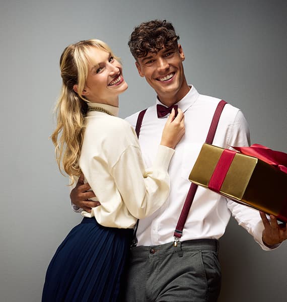 Couple souriant en tenue habillée, l’homme tenant un cadeau doré avec ruban rouge
