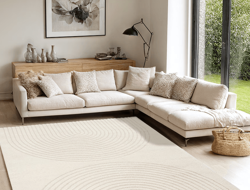 Beiger Ecksofa mit vielen Kissen auf großem Teppich, davor Korbtasche, dahinter Sideboard und Fenster zum Garten