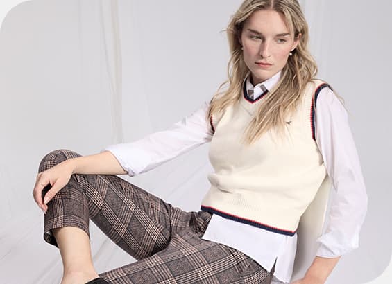 Frau trägt weiße Bluse, cremefarbenen Pullunder und karierte Hose, sitzt auf einem Stuhl und hat ein Bein ausgestreckt