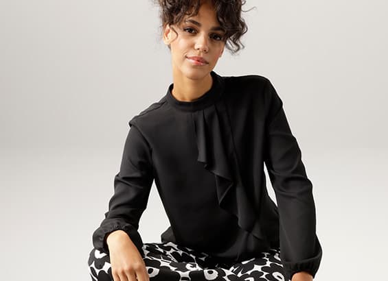 Femme assise en blouse noire à volants et pantalon noir à motifs blancs, face à l'objectif