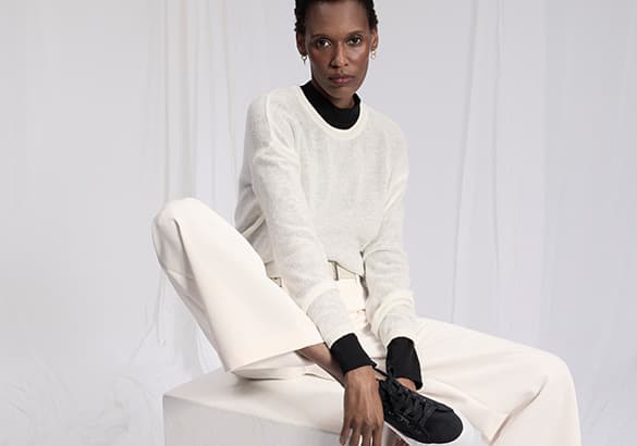 Personne assise sur un cube, portant un pull blanc et un pantalon blanc ample, avec des chaussures noires.