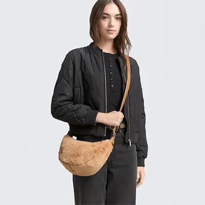 Frau in schwarzer Jacke und Hose trägt quer über der Schulter eine beige, bauchige Umhängetasche