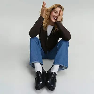 Person sitzt mit überschlagenen Beinen am Boden, trägt braune Jacke, blaue Jeans, weiße Socken und schwarze Schuhe