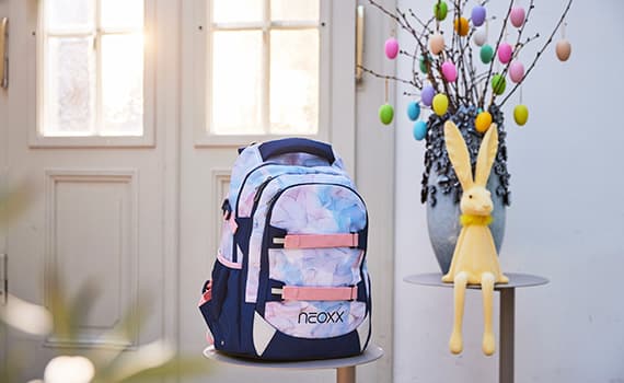 Schulrucksack mit buntem Blumenmuster neben Osterdekoration mit gelbem Hasen und bunten Eiern auf Zweigen.