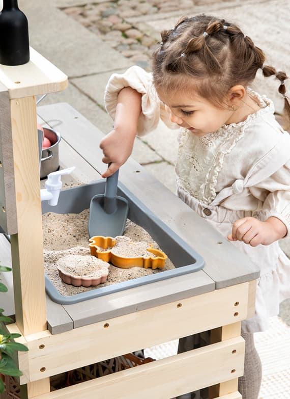 Kind spielt mit Sand und Schaufel an einem Holztisch mit Formen und Wasserhahn.