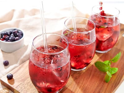 Drei Gläser roter Beerenlimonade mit Eiswürfeln und Strohhalmen auf Holzbrett, daneben frische Beeren und Minze.