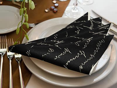 Schwarze Servietten mit weißer Schrift auf einem gedeckten Teller, daneben Besteck und Weingläser.