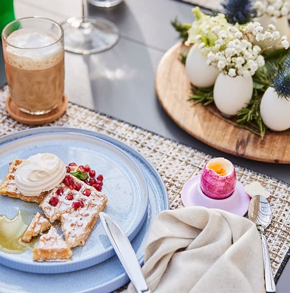 Waffeln mit Sahne und Granatapfel auf blauem Teller, daneben Kaffee, aufgeschnittenes Ei und Blumenarrangement in Eierschalen.