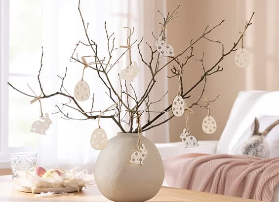 Décorations de Pâques en forme d'œufs et de lapins suspendues à des branches dans un vase beige sur une table
