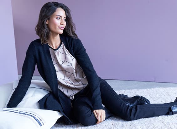 Personne assise sur un tapis, portant un cardigan noir et un haut à motif géométrique devant un mur violet.