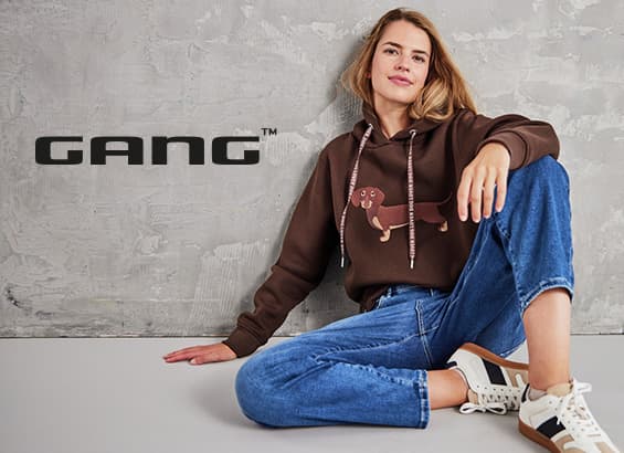 GANG Logo – sitzende Person in braunem Hoodie mit Dackelmotiv, blauer Jeans und Sneakers vor grauer Wand