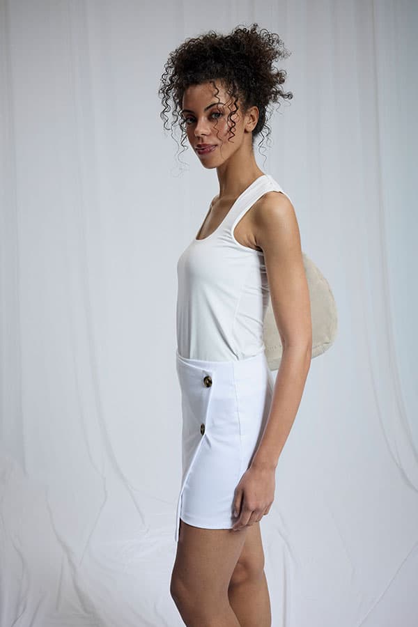 Personne debout de profil en débardeur blanc, jupe blanche boutonnée et sac à dos clair sur fond uni