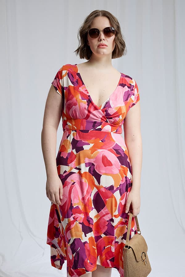 Robe portefeuille mi-longue à motifs abstraits rose, orange et violet, portée par une femme tenant un sac beige