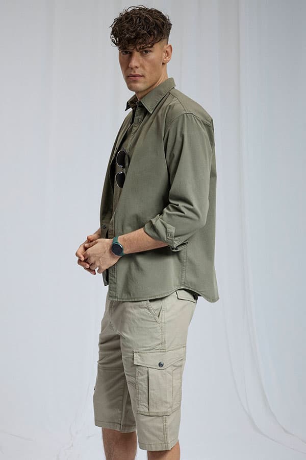 Homme de profil portant chemise kaki ouverte, short cargo beige à poches et montre, sur fond uni clair