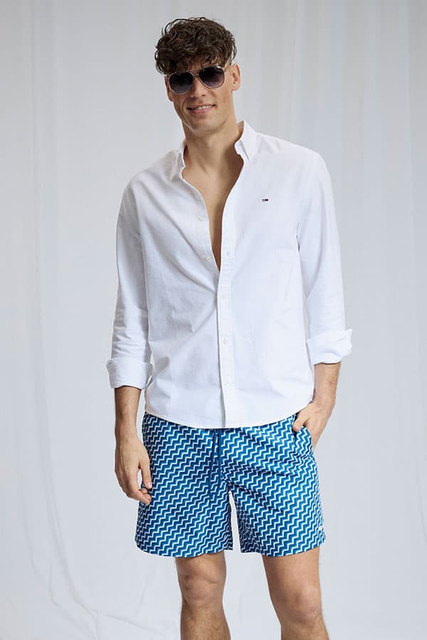 Mann in weißem Hemd und blauen Shorts mit Zickzackmuster steht vor hellem Hintergrund