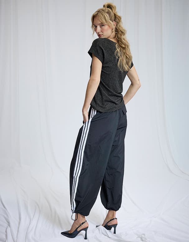 Personne debout de dos portant t-shirt noir, pantalon de survêtement noir à bandes blanches et escarpins à talons