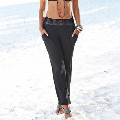 Pantalon de plage noir taille haute à ceinture ajourée, porté par une personne pieds nus sur le sable