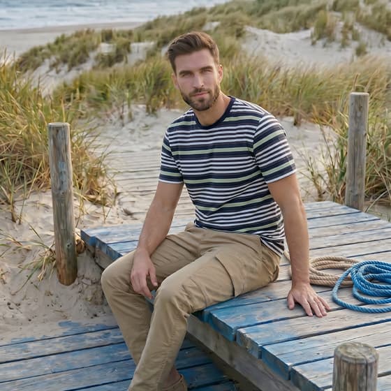 Mann in gestreiftem T-Shirt und Chinohose sitzt auf Holzsteg in den Dünen mit Blick zum Meer