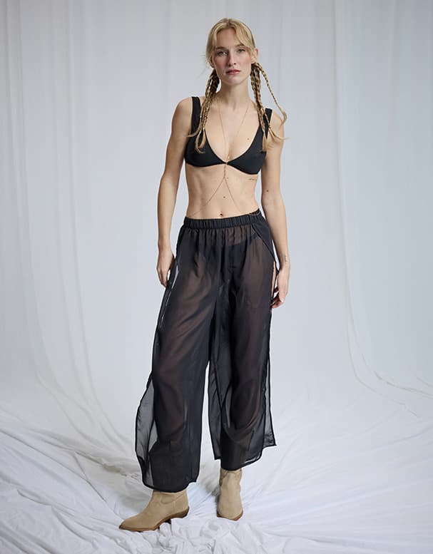 Frau mit geflochtenen Zöpfen trägt schwarzen Bikini-Top, weite transparente schwarze Hose und helle Stiefeletten im Studio