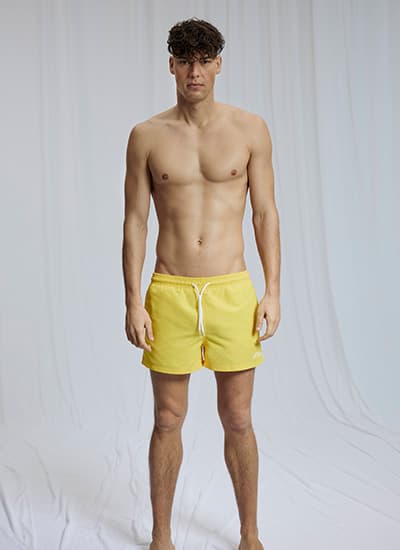 Mann steht frontal im Studio und trägt kurze gelbe Badeshorts mit weißem Kordelzug
