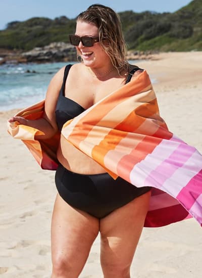 Frau im schwarzen Bikini am Strand, hält ein bunt gestreiftes Strandtuch um Schultern und Hüfte