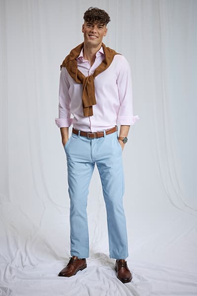 Personne debout en chemise rose, pantalon bleu clair, pull marron sur les épaules et chaussures habillées brunes