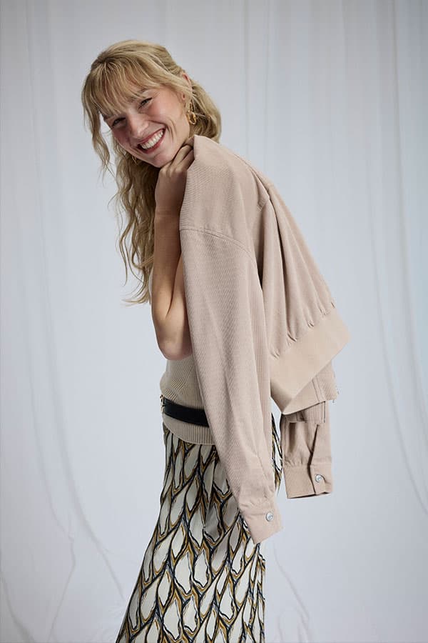 Personne souriante de profil, veste beige sur l’épaule, jupe longue à motif graphique noir et ocre