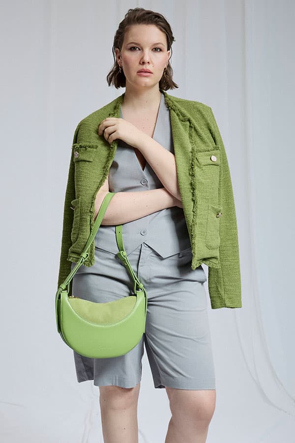 Femme debout en tailleur short gris, veste verte posée sur les épaules et sac bandoulière vert à la main