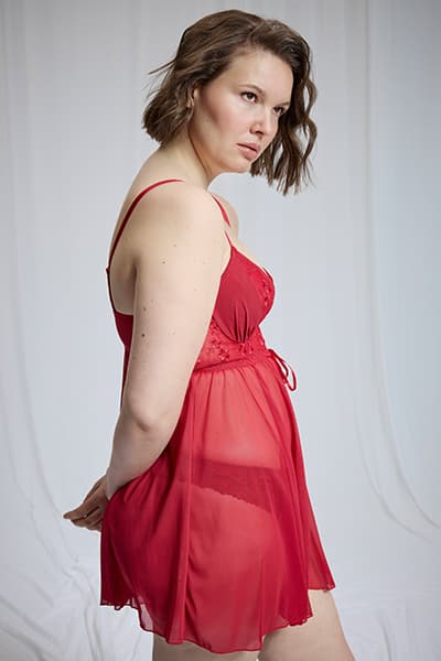 Femme de profil portant une nuisette rouge transparente avec bretelles fines, devant un fond drapé clair