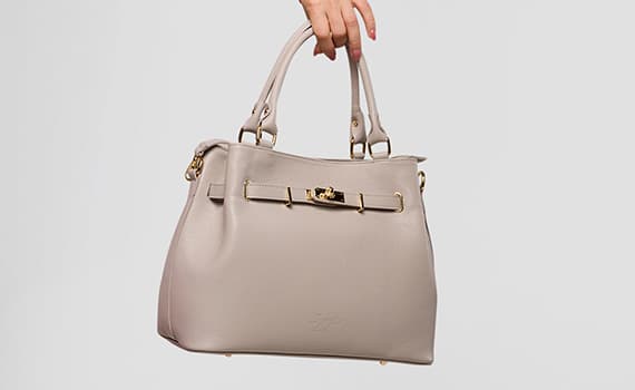 Hand hält beigefarbene Henkelhandtasche mit goldfarbener Schnalle vor hellem Hintergrund