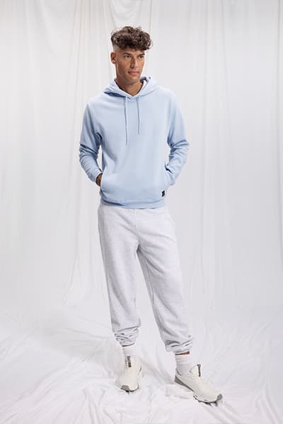 Mann in hellblauem Hoodie und grauen Jogginghosen, weiße Turnschuhe, Hände in Hosentaschen, weißer Hintergrund