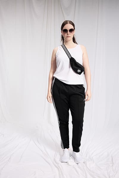 Frau trägt weißes ärmelloses Top, schwarze Jogginghose mit Streifen und schwarze Gürteltasche, steht vor weißem Hintergrund