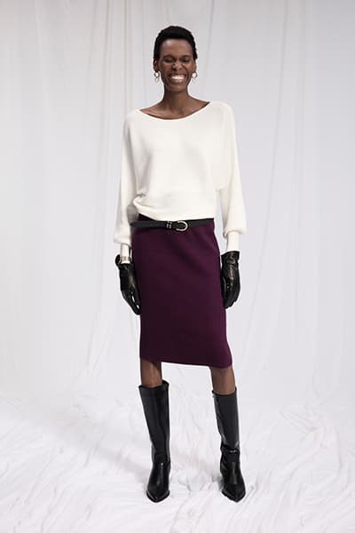 Frau trägt weißes Oberteil, lila Rock mit Gürtel, schwarze Stiefel und Handschuhe vor weißem Hintergrund