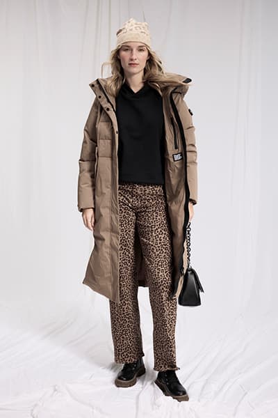 Frau trägt langen braunen Mantel, schwarze Bluse und Leopardenhose, hält schwarze Handtasche.