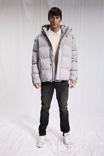 Mann trägt graue Daunenjacke über weißem Pullover, schwarze Jeans und weiße Sneakers vor weißem Hintergrund