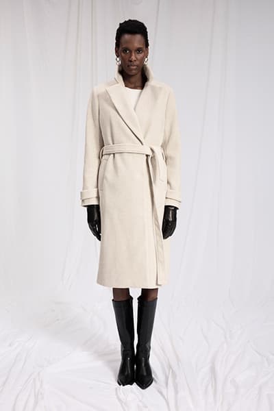 Frau trägt langen cremefarbenen Mantel mit Gürtel, schwarze Handschuhe und schwarze Stiefel. Gesicht ist unscharf.