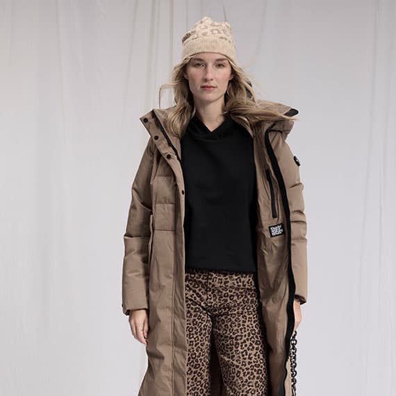 Frau trägt braunen langen Wintermantel, schwarzes Oberteil, Leopardenmuster-Hose und beige Mütze.